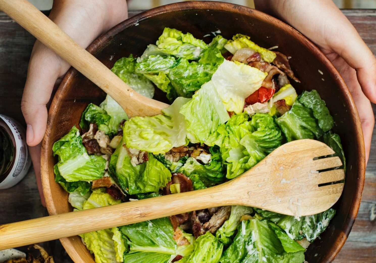 A person holding wooden spoons over a bowl of salad.